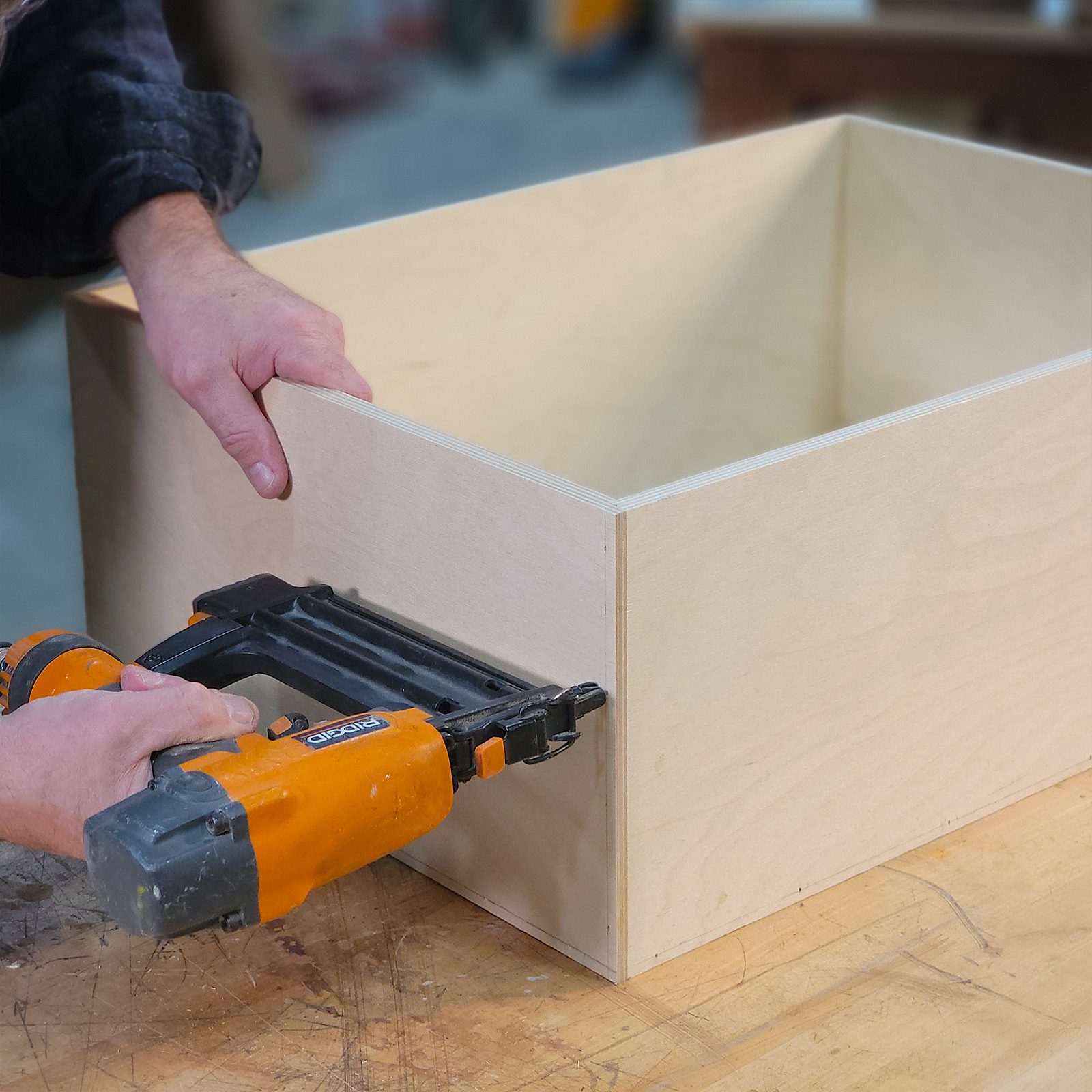 A person uses a nail gun to attach two pieces of plywood while working on a wooden surface in a workshop environment.