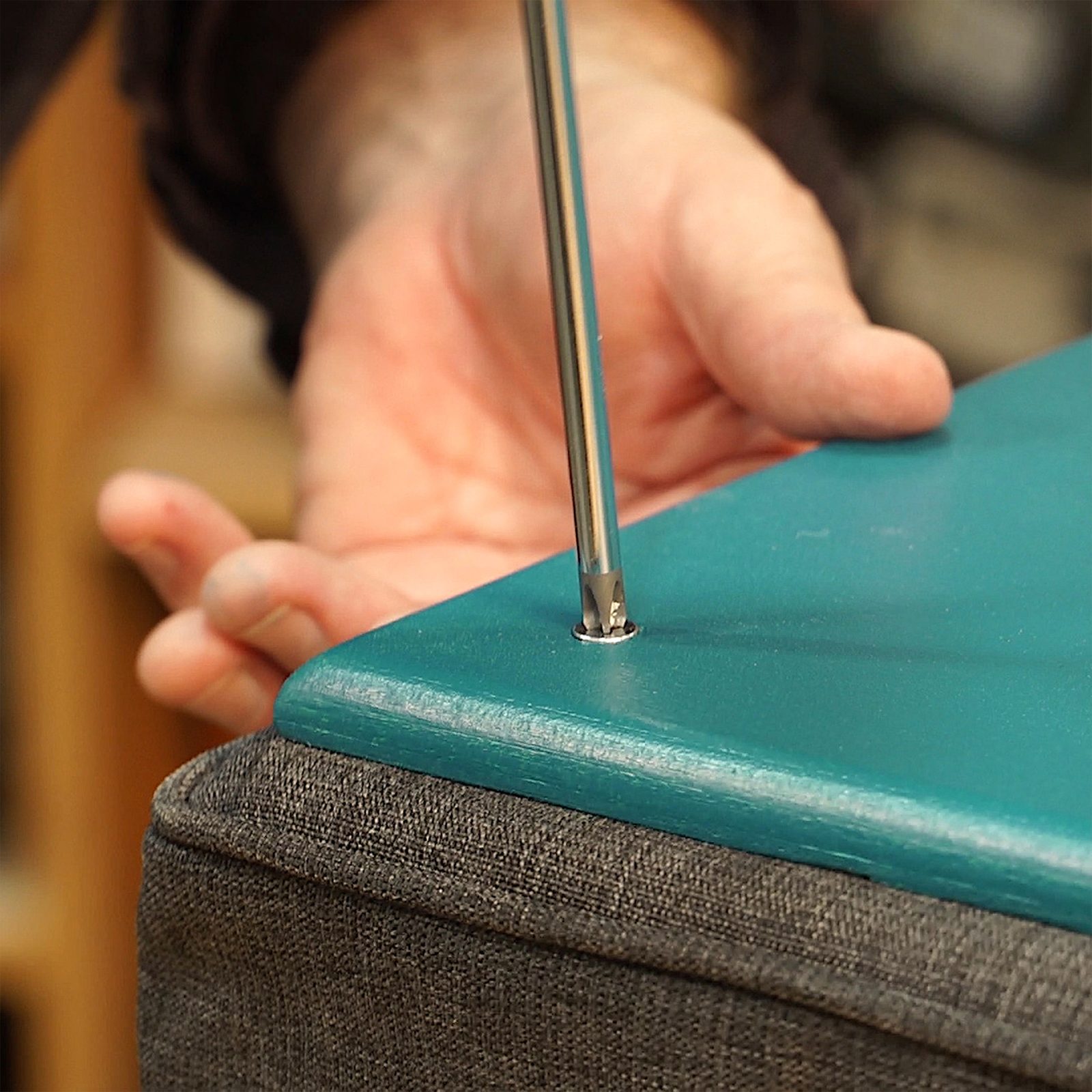 A hand grasps a tool, tightening it into a hole on a teal wooden surface, which rests on a gray fabric structure.