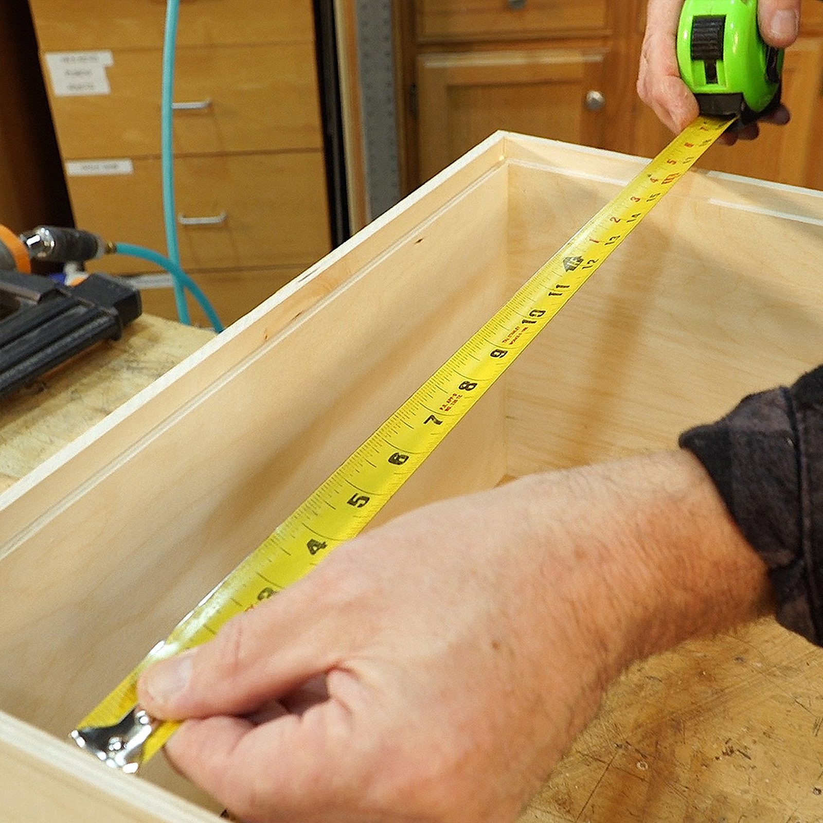 A hand holds a tape measure, extending it inside a wooden box. The setting features tools and wooden surfaces, suggesting a workshop environment.