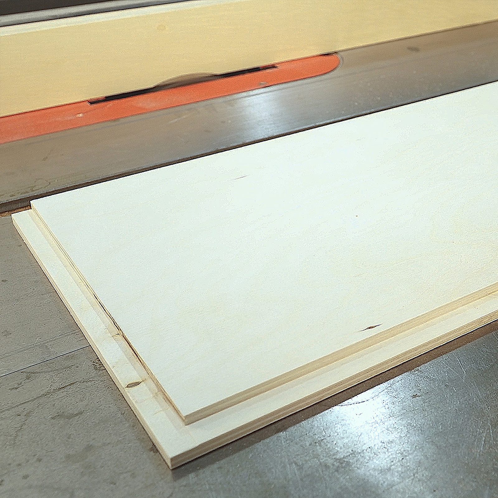 Two wooden plywood sheets lie flat on a table saw surface, ready for cutting in a workshop setting with a visible cutting blade and control panel.