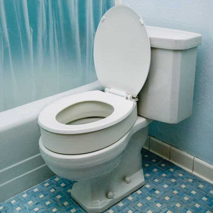 A white toilet sits in a bathroom, its lid open, alongside a pale blue wall and a textured floor, creating a clean, functional space.