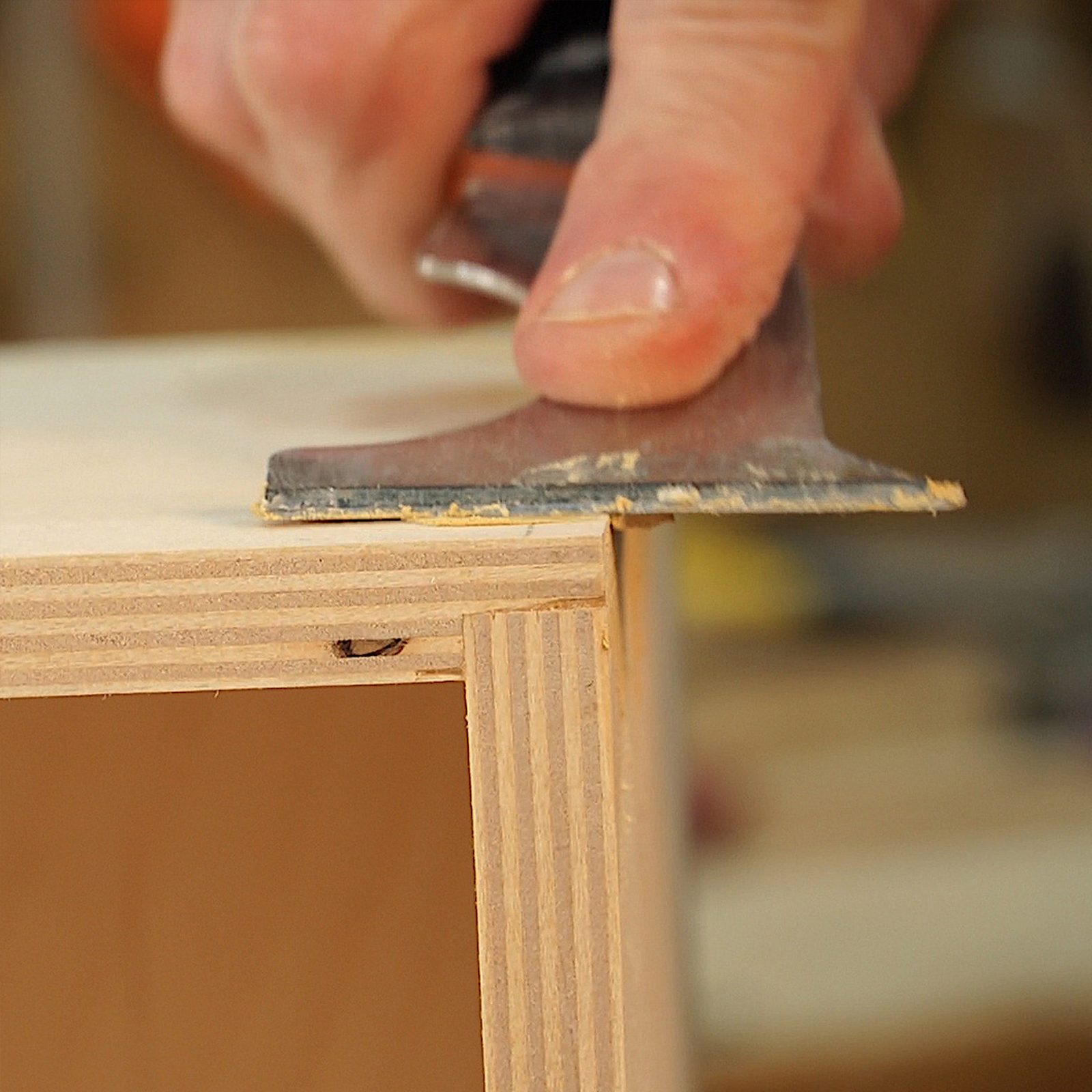A hand holds a scraping tool, smoothing the edge of a wooden surface, surrounded by workshop equipment and materials, indicating a woodworking project.