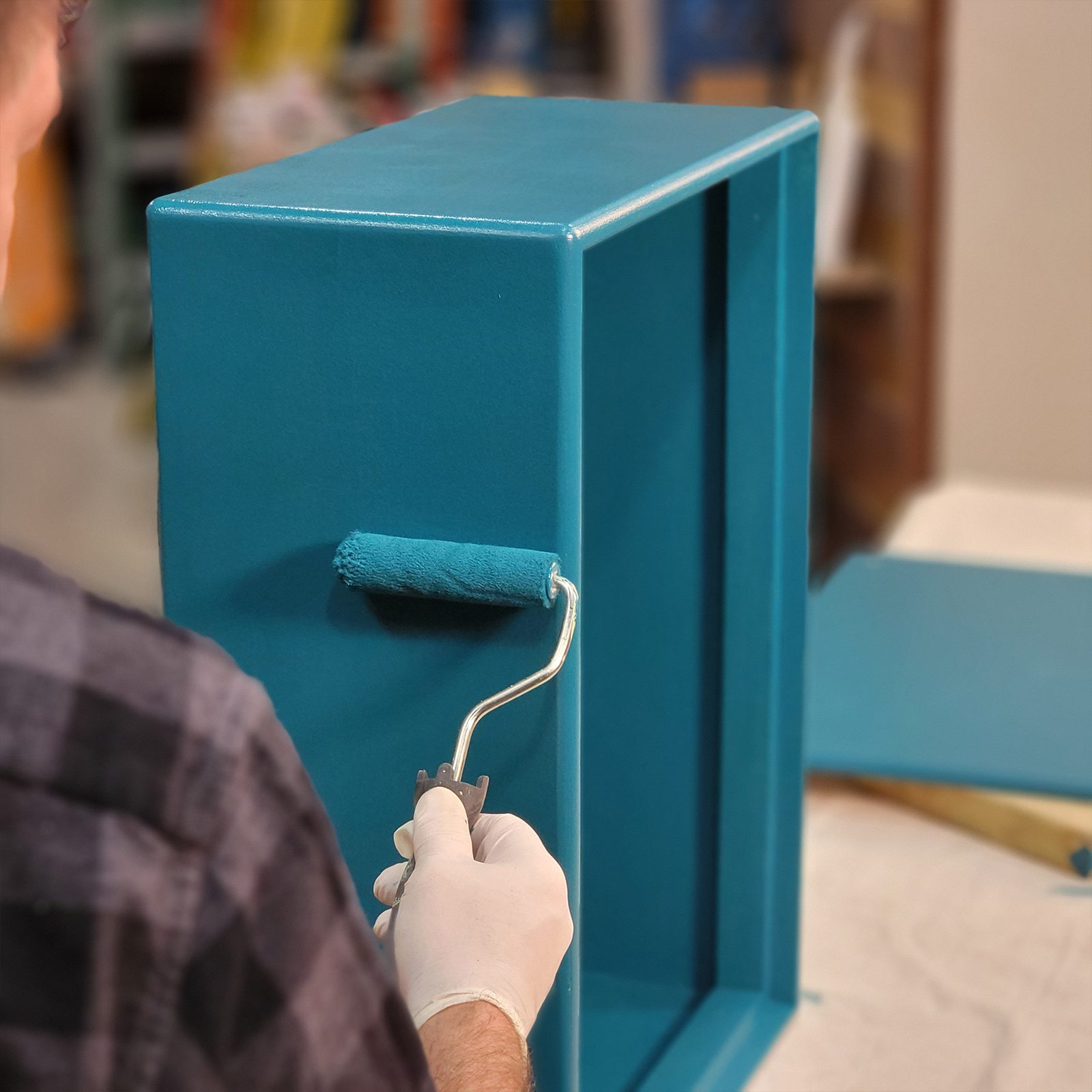 A hand in a glove rolls paint onto a teal cabinet in a workshop, with a partially painted surface and tools in the blurred background.