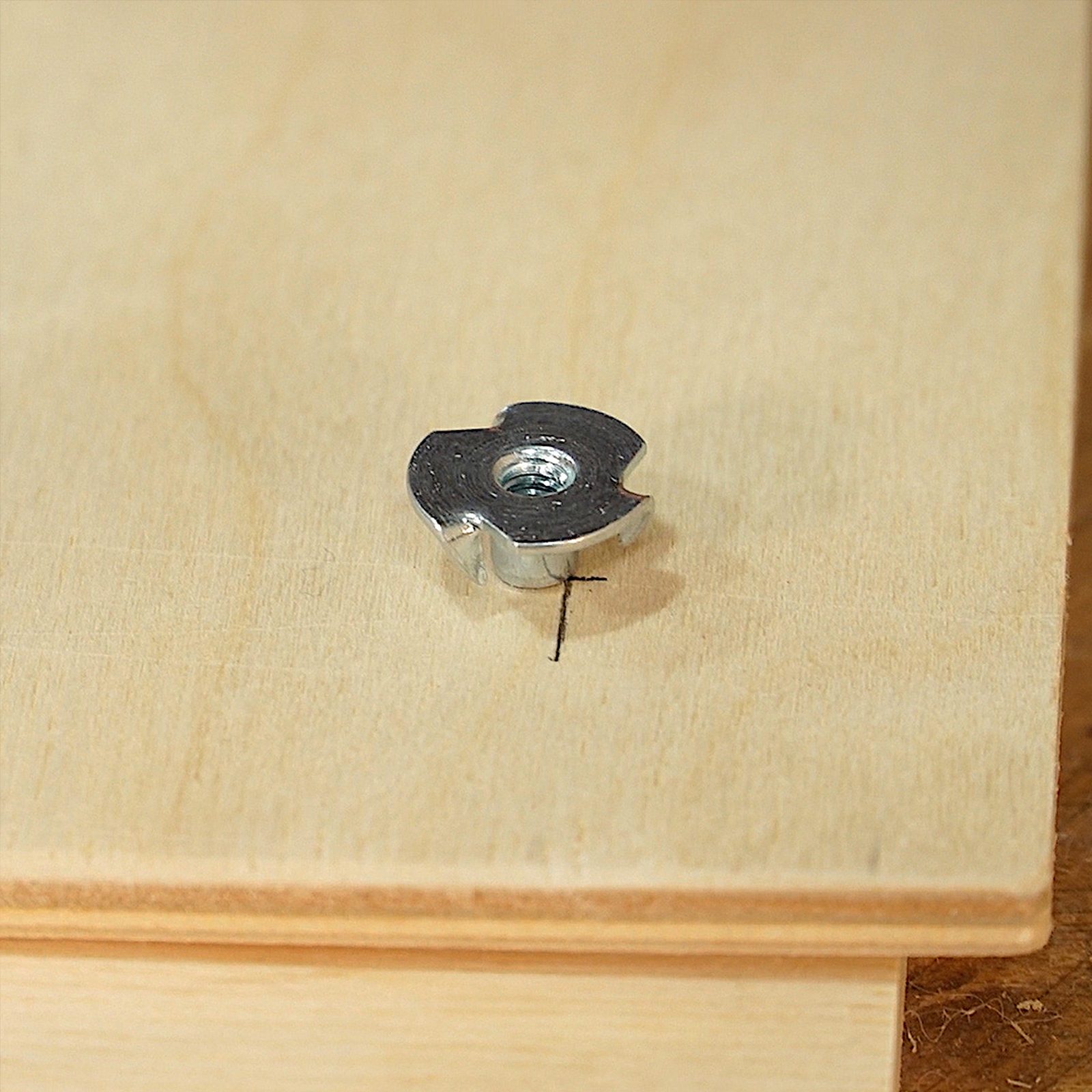 A metallic fastener rests on a wooden surface, aligned with a black marking, indicating a precise location for installation or drilling.