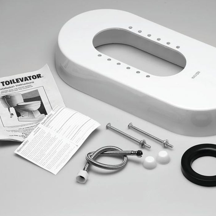 A white toilet riser piece lies on a flat surface with installation instructions, screws, a flexible hose, and rubber fittings arranged around it.