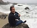 A person sits on a rock near the shore, smiling as waves crash against the beach under a cloudy sky.