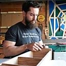 A man with a beard works on a wooden drawer in a workshop, focusing intently on his task amidst various tools and colorful art pieces.