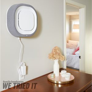 A wall-mounted air purifier operates with a power cord, positioned above a wooden dresser displaying a vase with dried flowers and decorative candles.