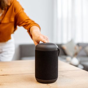 A woman touches a black speaker on a wooden table, while holding a phone. A cozy living room with a gray couch and decorative pillows is in the background.