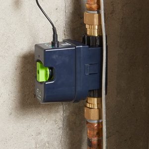 A device is mounted on a wall near copper pipes, indicating a water monitoring system. It's connected to a power source with a wire.