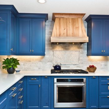 Blue kitchen cabinets with a modern design complement a white countertop and stainless steel appliances, illuminated by warm lighting against a stone backsplash.