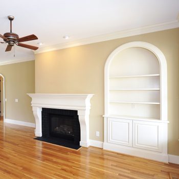 A ceiling fan spins above a wooden floor, while a white fireplace and built-in shelves enhance the light-colored walls of a spacious living room.