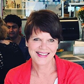 A woman with short hair, smiling, wears a red jacket. In the background, a café environment with blurred figures and a coffee machine is visible.
