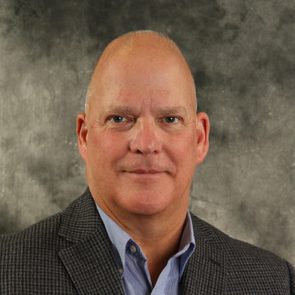 A man with a bald head and slight stubble, wearing a checkered blazer over a blue shirt, gazes directly at the camera against a textured gray background.