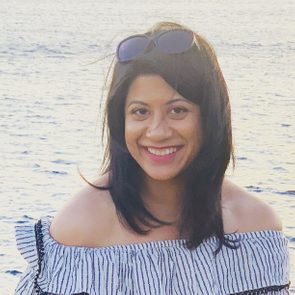 A woman with shoulder-length hair smiles while wearing an off-shoulder striped top, standing in front of a shimmering body of water.