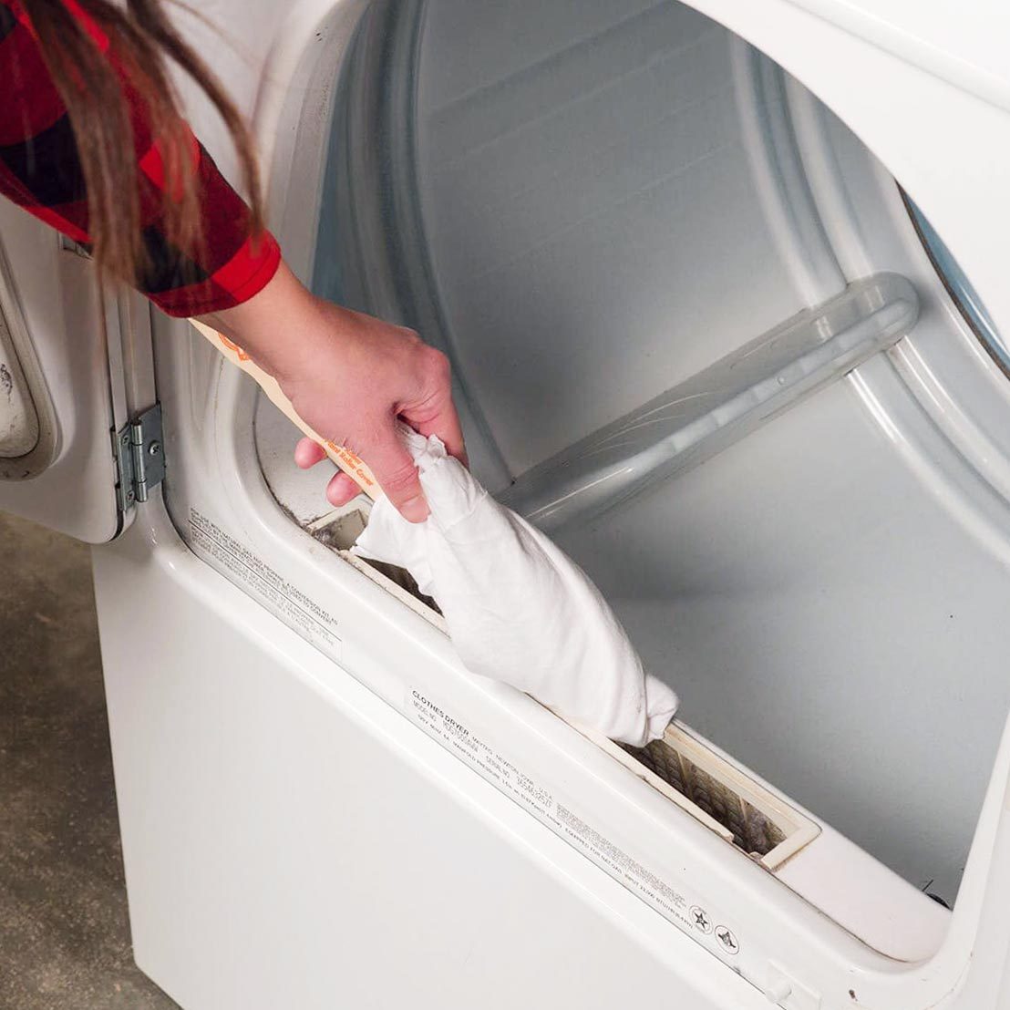 A person removes a white lint trap from a clothes dryer, located in a domestic environment with a gray floor and clean interior.