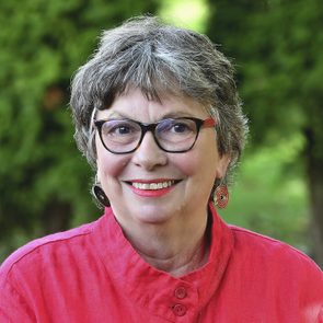 A smiling older woman with short gray hair wears glasses and colorful earrings, standing outdoors against a backdrop of green foliage.