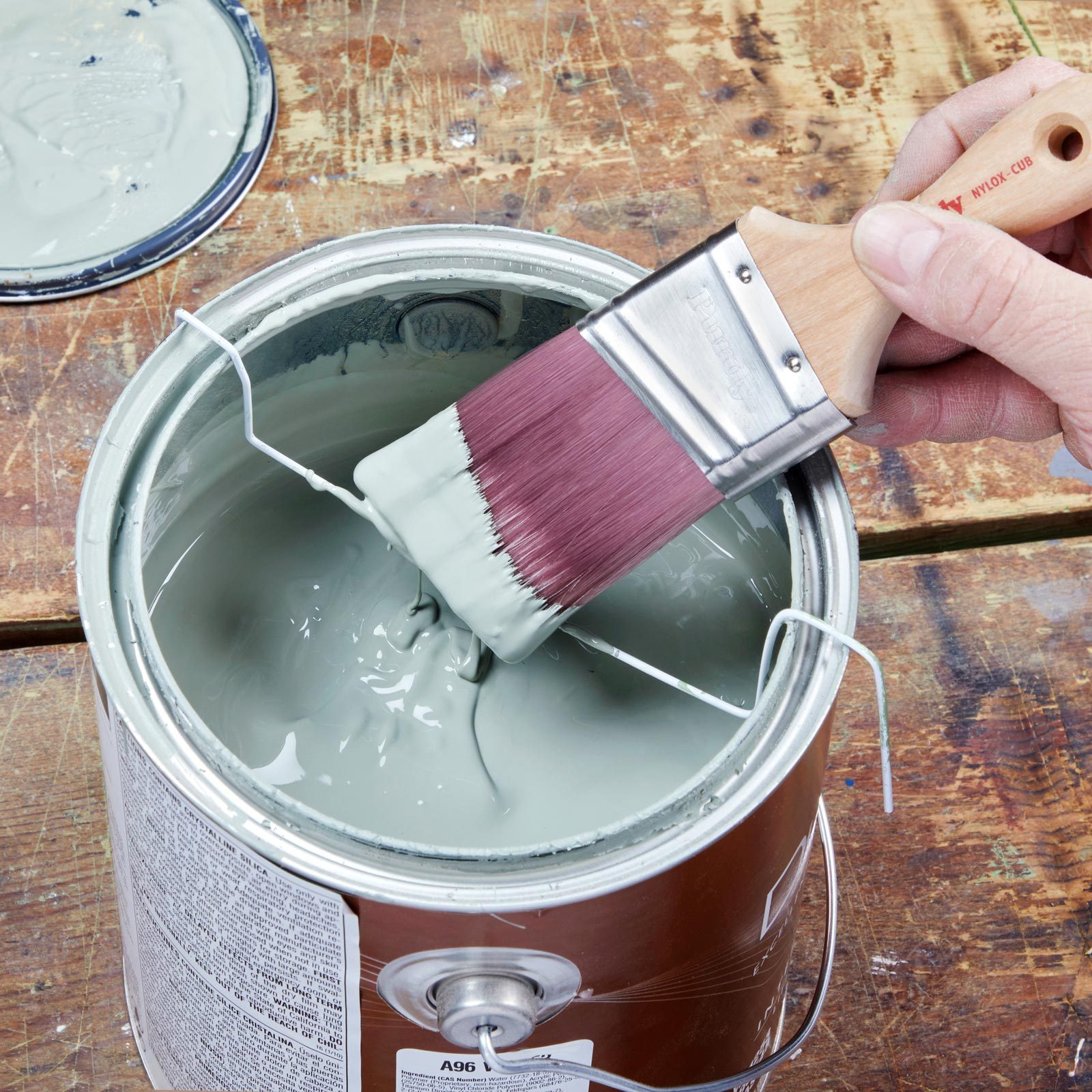 A paintbrush lifts green paint from a metal can, ready for application, set against a wooden surface with a rustic appearance.