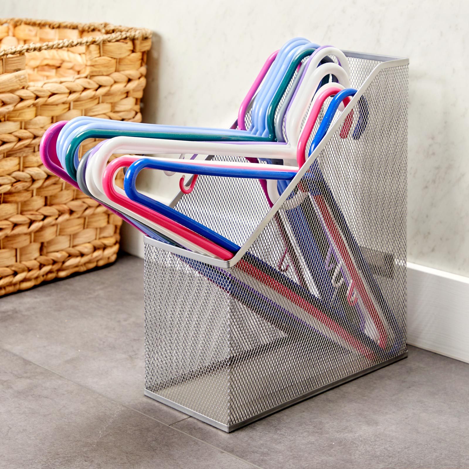 A wire mesh holder displays colorful plastic hangers, organized vertically. It's situated beside a woven basket in a well-lit indoor space with gray flooring.