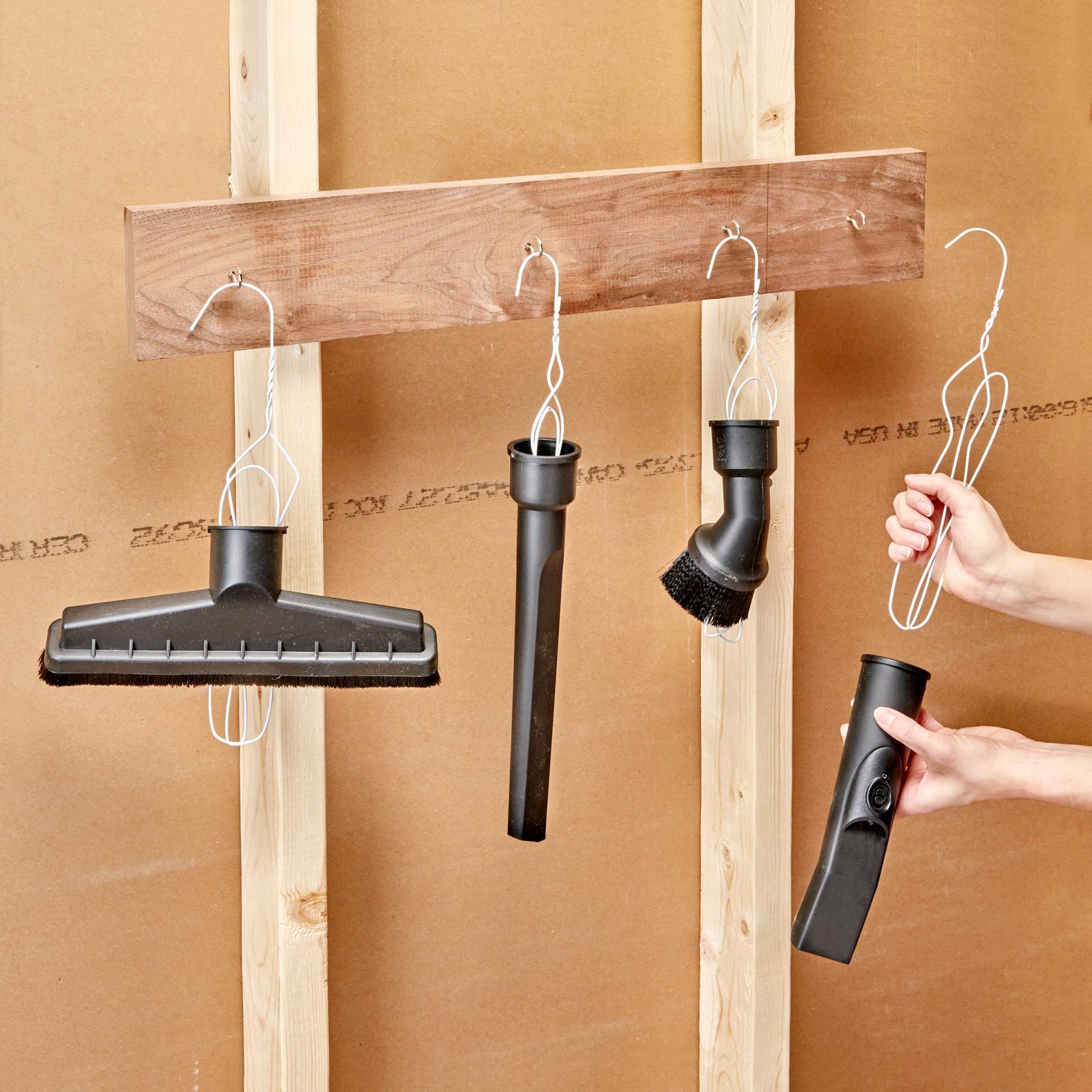 A wooden board with four black cleaning tool attachments hangs on a wall, while a hand holds a wire hook for one of the tools.