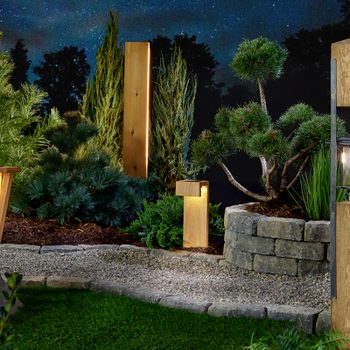 Stylish outdoor lights illuminate a landscaped garden at night, surrounded by various greenery and stone features, creating a serene ambiance.