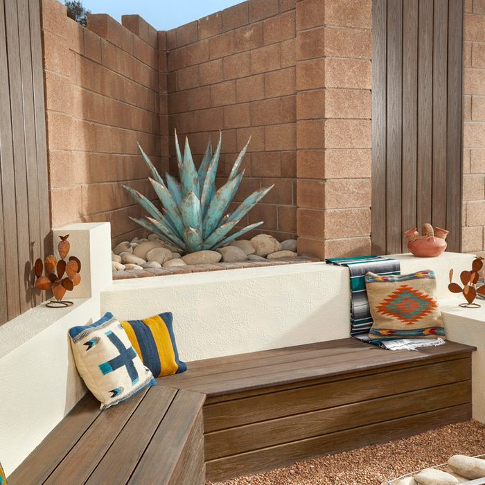 A blue-green agave plant grows among stones in a cozy outdoor nook, flanked by wooden walls and decorated with colorful pillows and pottery.