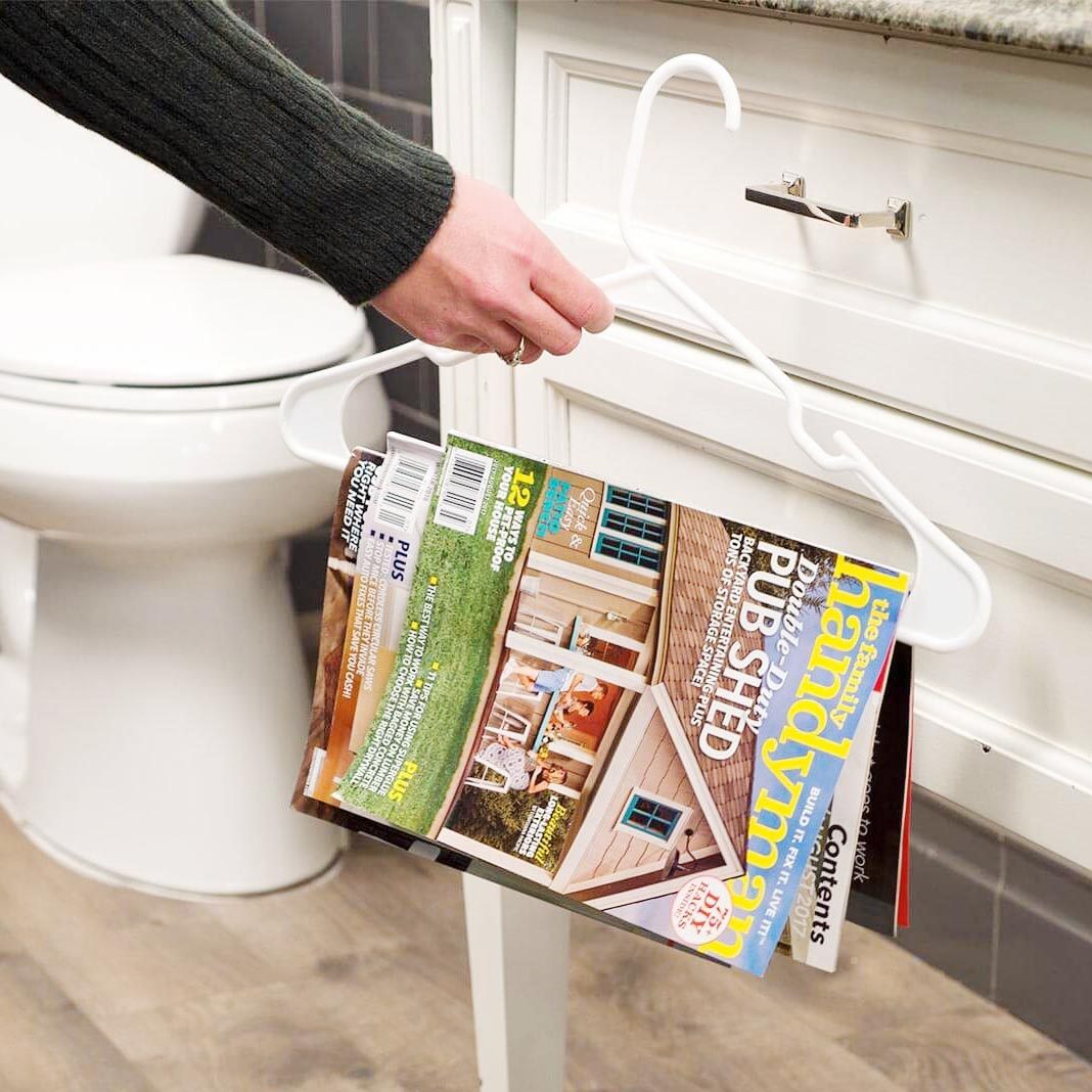A hand holds a white hanger with magazines hanging from it, positioned next to a toilet in a bathroom with wooden flooring.