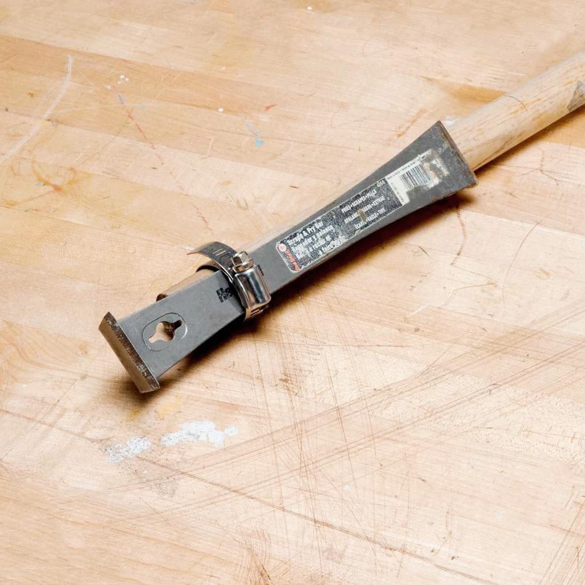 A metal scraper with a wooden handle lies on a textured wooden surface, appearing worn and attached with a metal clamp.