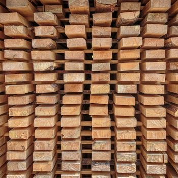 Stacks of wooden planks are neatly arranged, lying horizontally and vertically, showcasing varied textures and colors in a structured, orderly manner.