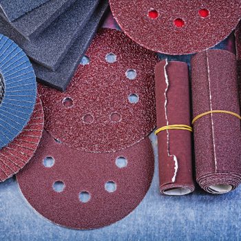 Various sanding materials, including discs and sheets, are stacked and arranged on a textured blue surface, demonstrating their diverse shapes and sizes for surface finishing.