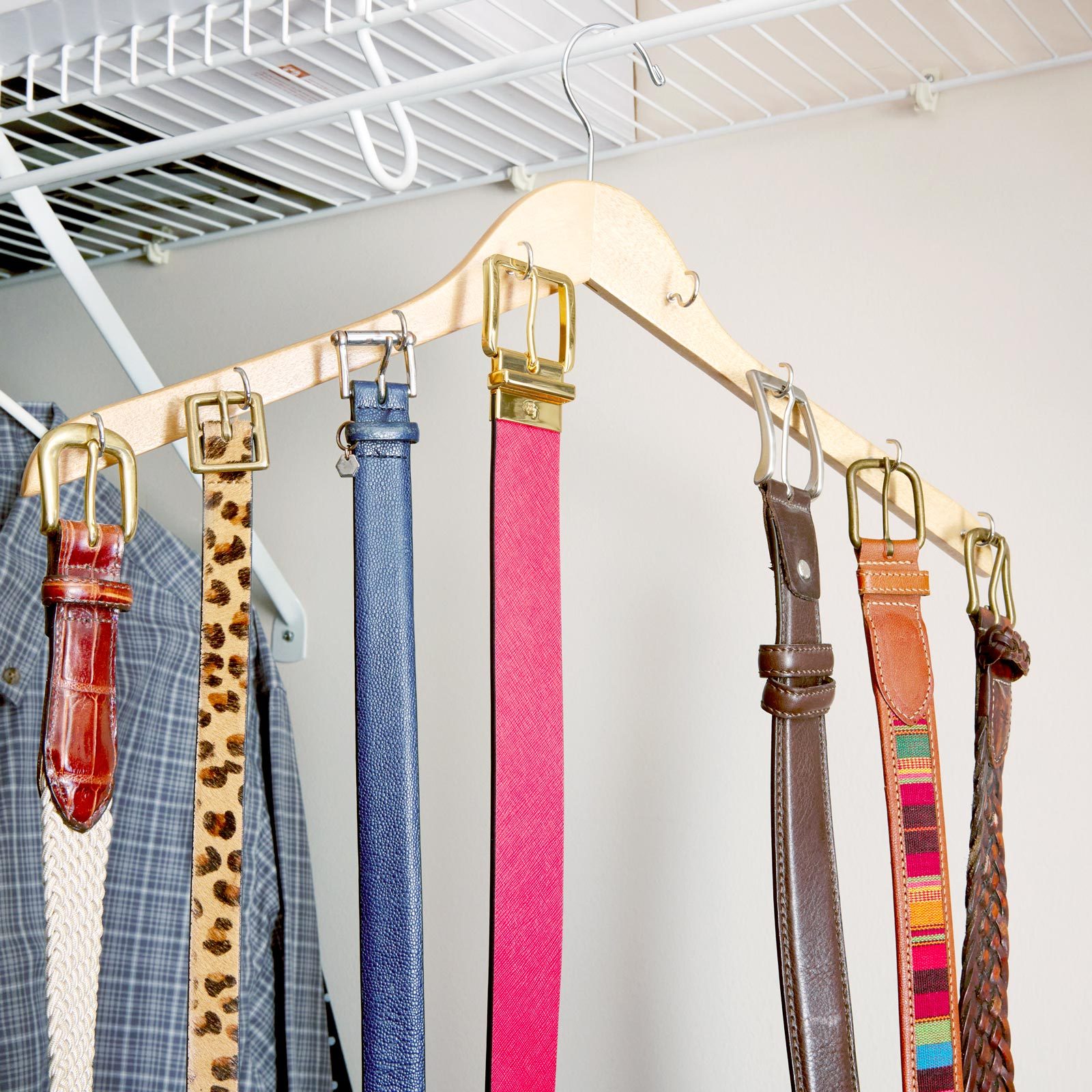A wooden hanger holds various belts, each displaying distinct colors and patterns, organized neatly against a neutral wall, with a shirt hanging nearby.
