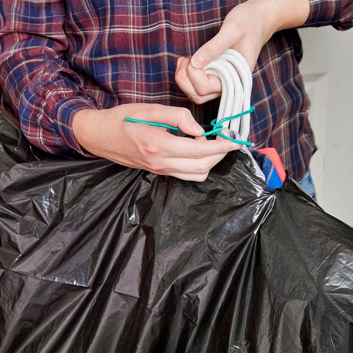 A person ties a black garbage bag closed using a green tie, holding the bag with one hand while manipulating the tie with the other.