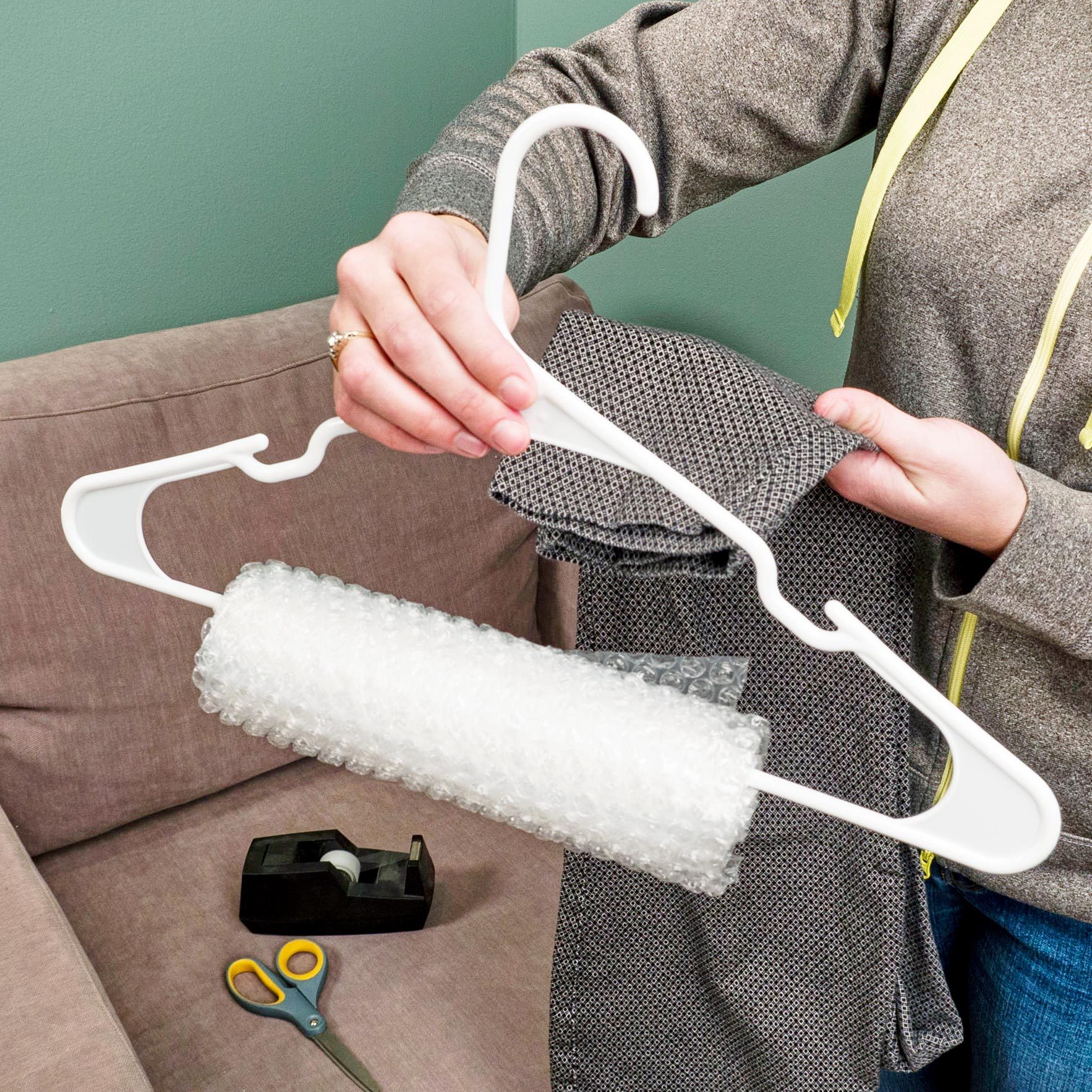 A person holds a hanger with bubble wrap while preparing to drape a patterned fabric, sitting on a couch near a tape dispenser and scissors.