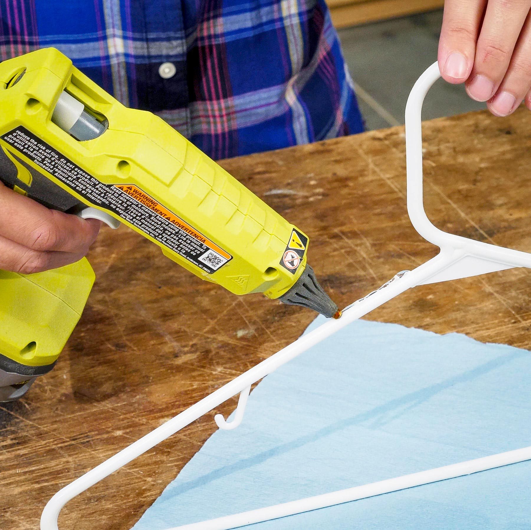 A hand holds a yellow glue gun, applying adhesive to a white hanger on a wooden surface, with a blue cloth underneath.