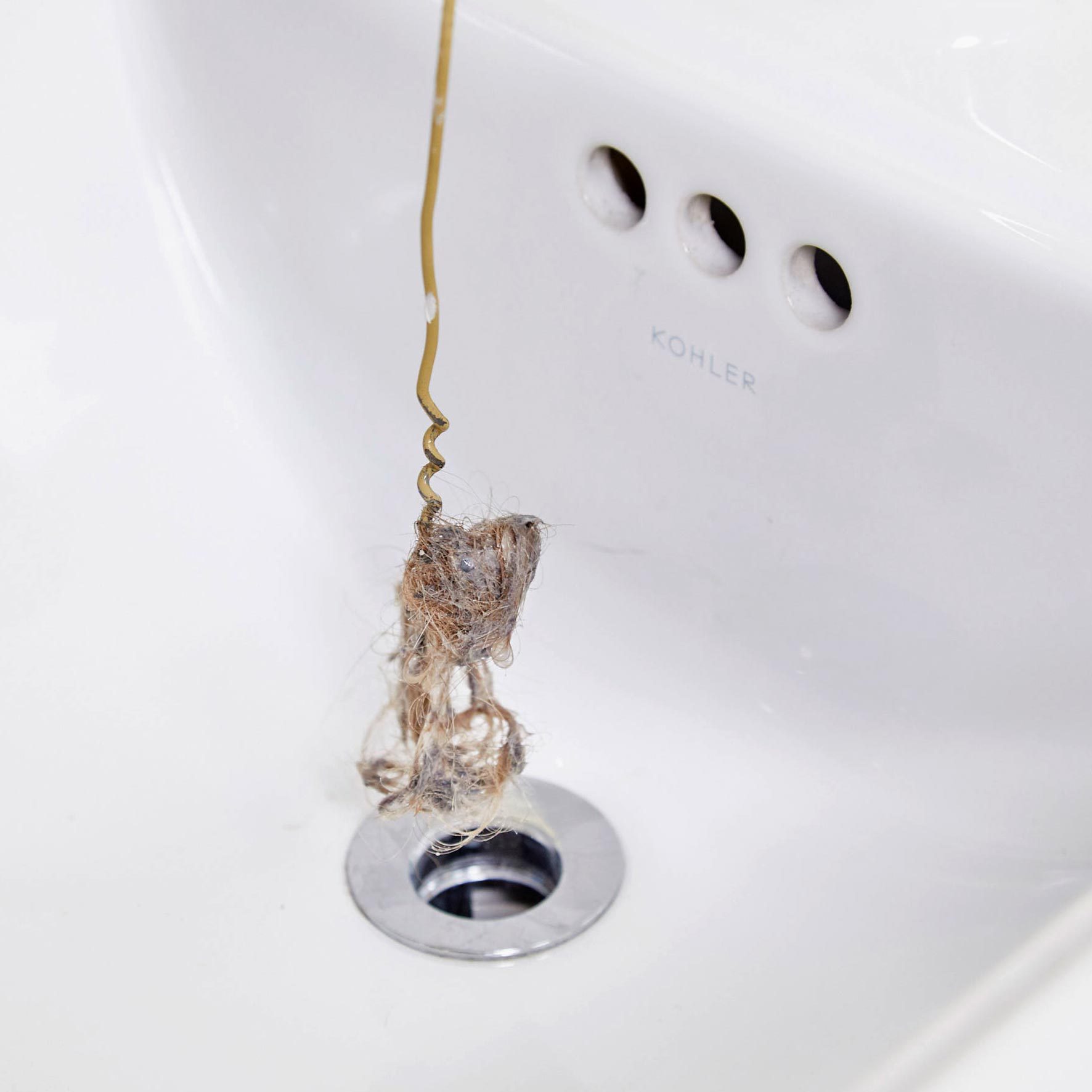 A plumbing snake extracts a tangled clump of hair and debris from a sink drain, positioned next to a polished chrome drain cover in a white basin.