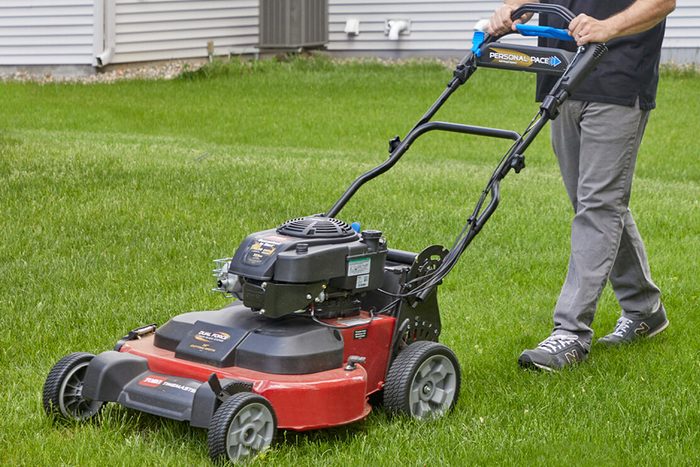 A man uses a lawn mower to cut grass in a backyard, surrounded by a home and landscaping features, under clear conditions.