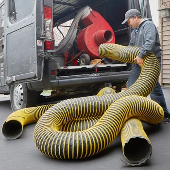 A man lifts a long yellow and black hose from a van, moving it toward a red machine inside, set on a driveway near a house.