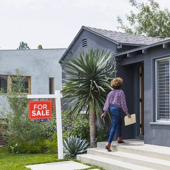 A woman carrying documents walks up steps towards a gray house, passing a "FOR SALE" sign in a landscaped yard with green plants.