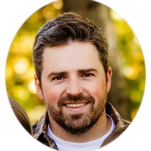A man with a beard smiles, wearing a plaid shirt, surrounded by blurred greenery, suggesting an outdoor setting.