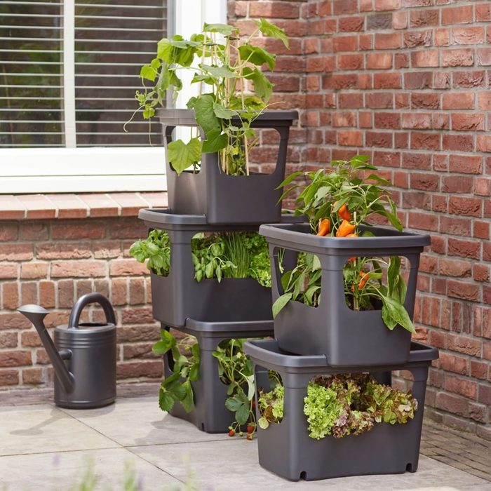 Stacked planter boxes filled with various plants, including herbs and vegetables, are positioned near a brick wall, alongside a watering can on a stone surface.