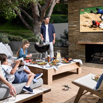 A family enjoys a barbecue on a patio, with a man grilling, children eating, and a sports game displayed on a wall-mounted TV.