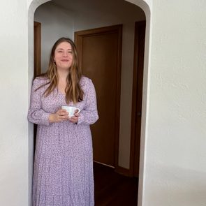 A woman in a floral dress stands in a doorway holding a cup, smiling in a well-lit room with wooden floors and brown doors.