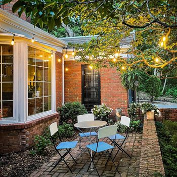 A table with four chairs is set outside a brick house, illuminated by string lights among leafy trees, creating a cozy atmosphere.