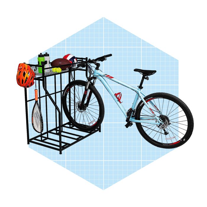 A mountain bike rests on a black stand, holding sports equipment like helmets, balls, and a racquet, against a light blue grid background.