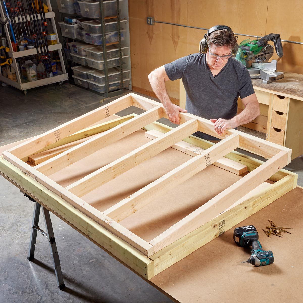 A person wearing safety glasses assembles a wooden frame on a worktable, surrounded by tools and storage containers in a workshop environment.