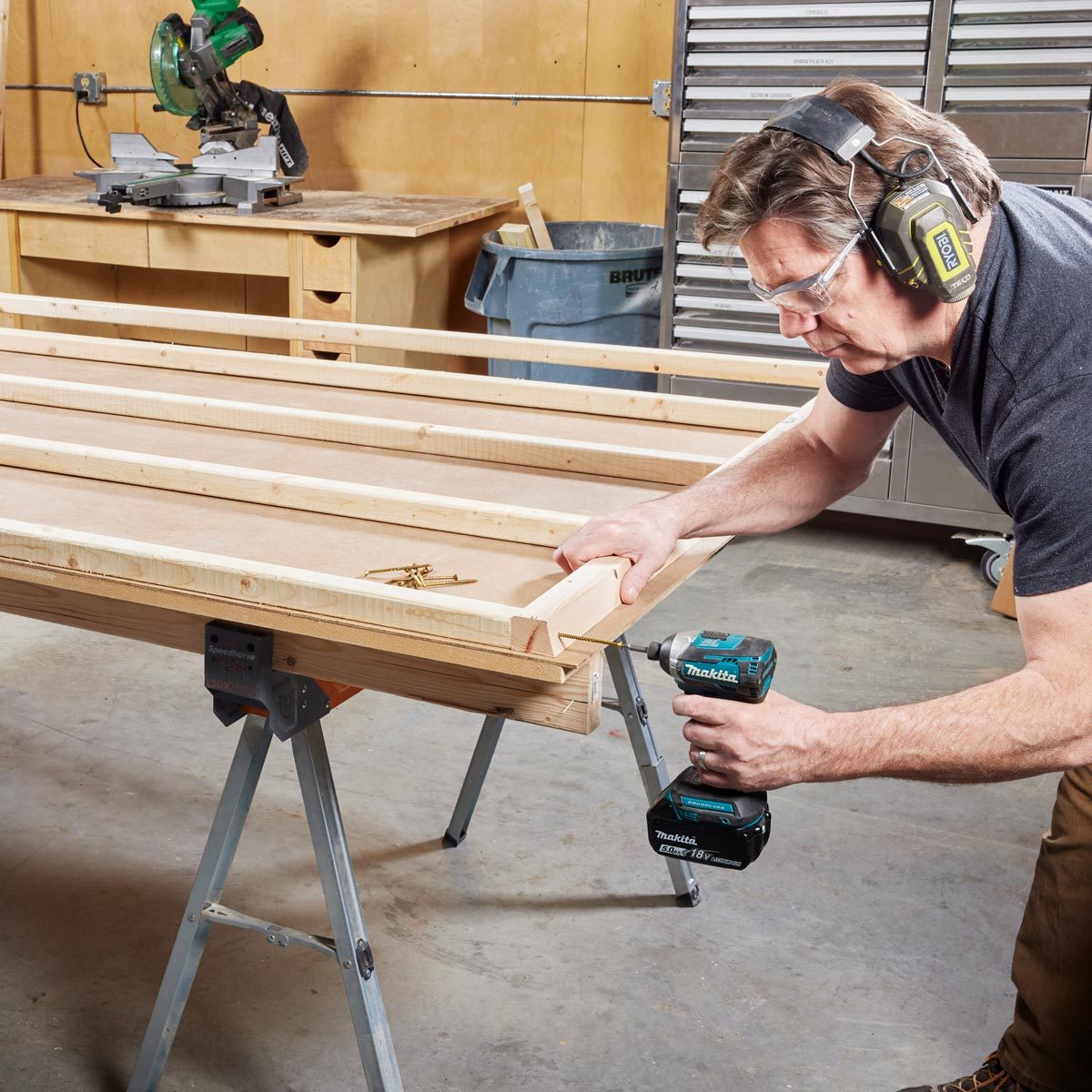 A man uses a power drill to secure wooden pieces on a workbench, surrounded by tools and a clean workshop environment.