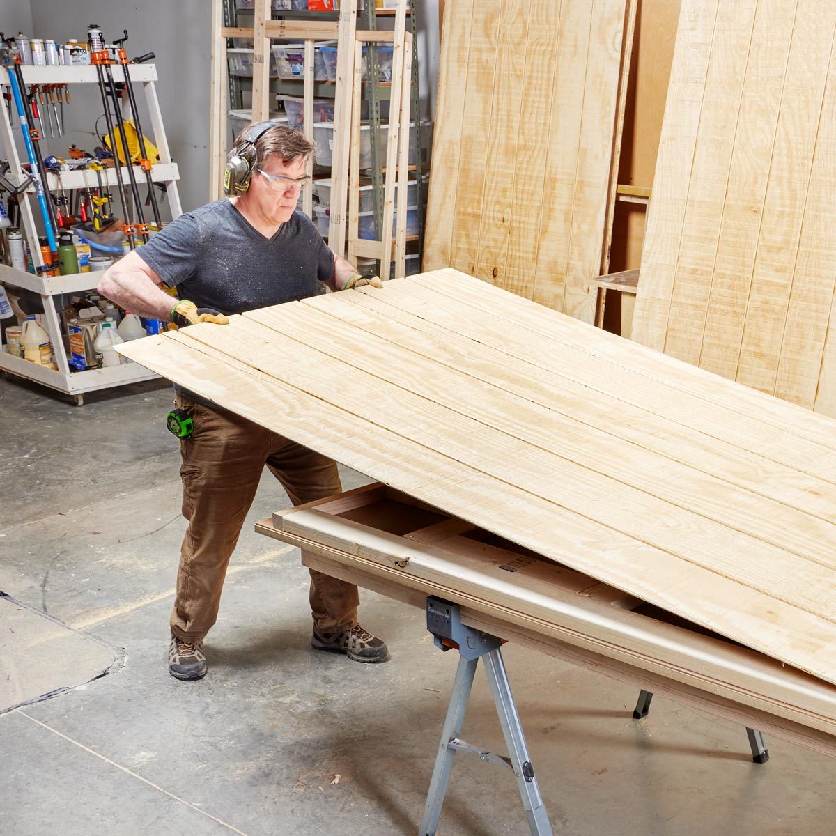 A man lifts a large wooden board while wearing gloves and headphones. The workshop is filled with tools and stacked plywood in the background.