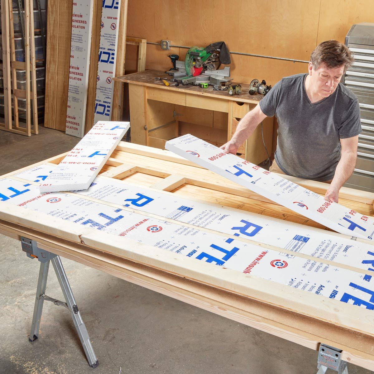 A man places insulation boards on a wooden frame located on a work table in a workshop filled with tools and materials.