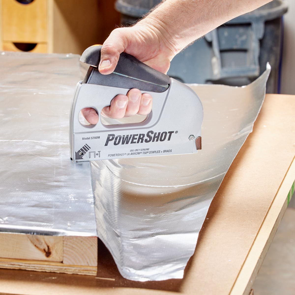A hand is using a stapler to secure aluminum foil to a wooden surface in a workspace, with tools and materials visible in the background.