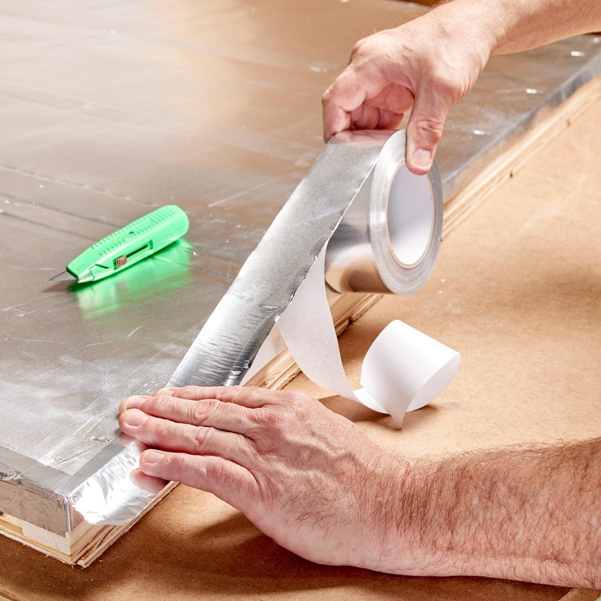 Hands apply silver tape onto a surface, with a utility knife and green cutter nearby on a wooden table, highlighting a DIY project.